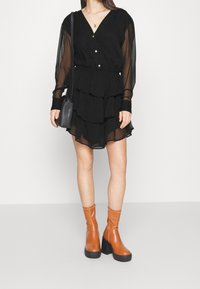 Žena v černých vrstvených mini šatech s průhlednými dlouhými rukávy, hnědými platformovými kotníkovými botami a malou černou kabelkou přes rameno.