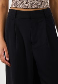 Μαύρο ταυτόχρονα παντελόνι διαθέτει υψηλή μέση, πιέτες, πλαϊνές τσέπες και απαλό υφάσμα. Το υλικό φαίνεται ελαφρύ και έχει ωραία drape.