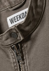 Κοντινή λήψη γκρι υφασμάτινου σακακιού με μεταλλικό φερμουάρ και κουμπί κοντά στον γιακά· η ετικέτα γράφει "WEEKDAY".