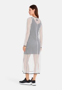 Langes, durchsichtiges Kleid mit einem schwarz-weißen Punkte-Muster, das einen hohen Kragen, lange Ärmel und schwarze Verzierungen am Saum aufweist, kombiniert mit schwarzen Sneakern.