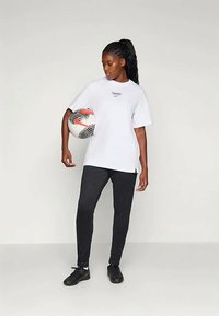 Balta T-krekls ar uzrakstu "PARIS", melni sporta bikses un melni futbolista apavi. Tur rokās krāsains futbols ar izteiktiem rakstiem.