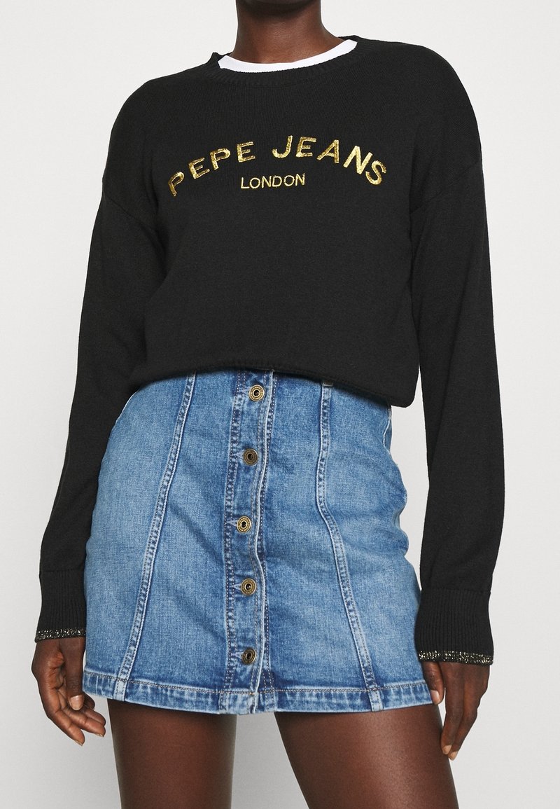 Maglione nero a maniche lunghe con ricamo dorato "PEPE JEANS LONDON", abbinato a una gonna di jeans blu con bottoni frontali e un taglio dritto.