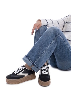 Sneakers bianco e nero con suola spessa color marrone chiaro, caratterizzati da un design slanciato e lacci bianchi, abbinati a jeans blu con risvolto.