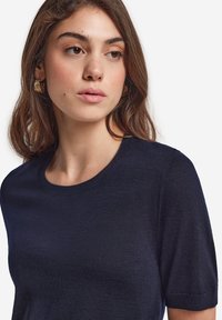 Navyblå topp med kort ärm och rund halsringning, gjord av lätt tyg. Modellen bär guldfärgade texturerade örhängen mot en enfärgad bakgrund.