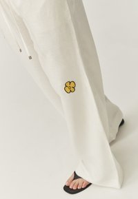 Witte, losse linnen broek met gele bloemenborduursel op het rechterbeen. Gecombineerd met zwarte sandalen, die een gladde textuur tonen.