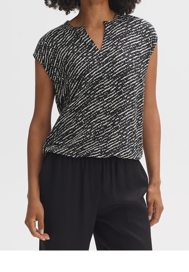 Black and white patterned short-sleeve top with a V-neck and relaxed fit, made from a lightweight fabric. Paired with black pants.