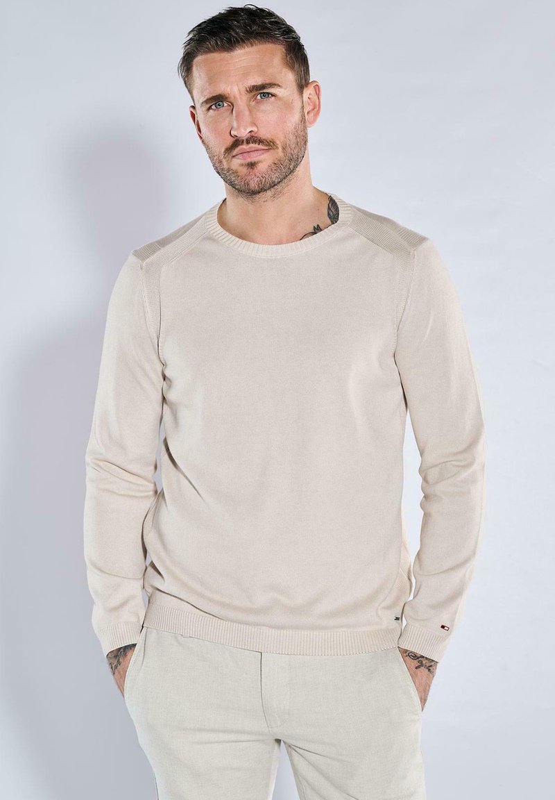 Beiger Strickpullover mit Rundhalsausschnitt, gerippten Bündchen und Schulterakzenten, kombiniert mit hellen Hosen. Glatte Textur über die gesamte Fläche.