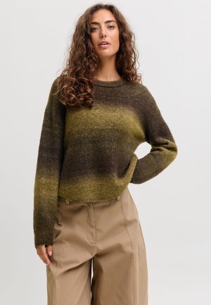 Femme aux cheveux bouclés portant un pull dégradé marron et vert et un pantalon large beige, debout devant un fond clair uni.