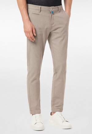 Homme portant un pantalon beige slim et des baskets blanches, debout avec une main dans la poche sur un fond uni.