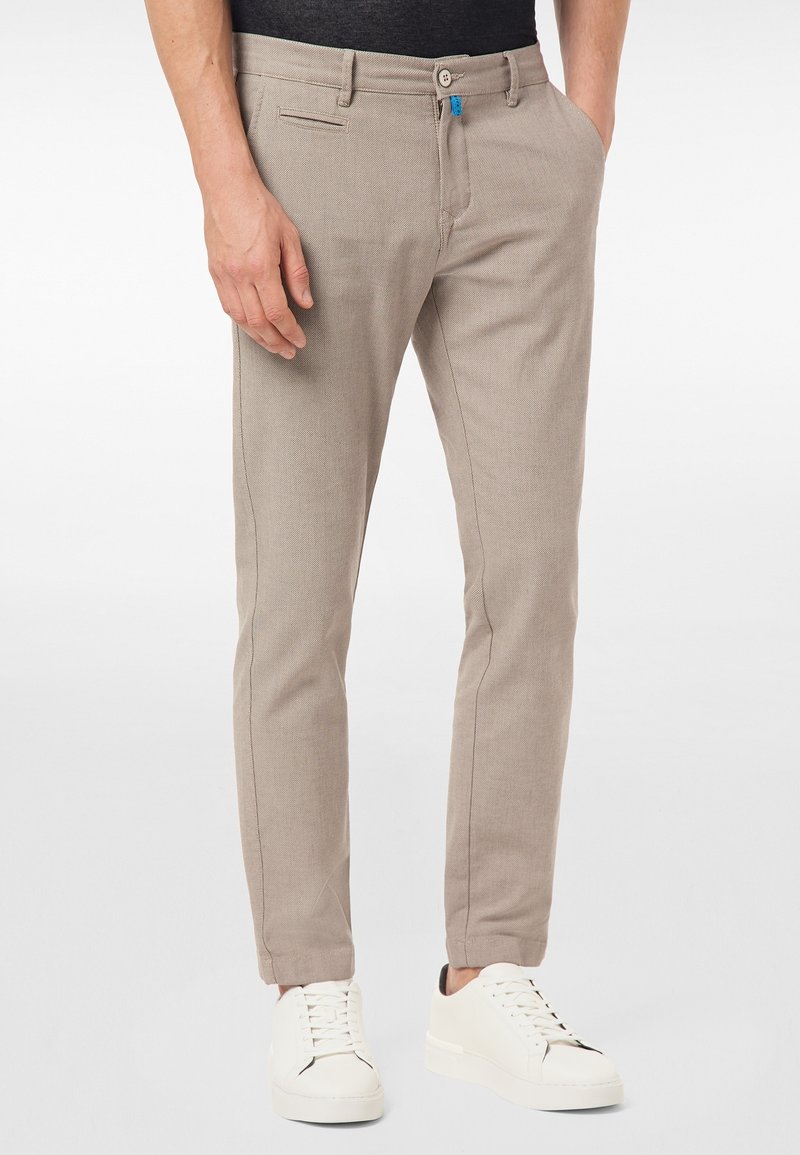 Homme portant un pantalon beige slim et des baskets blanches, debout avec une main dans la poche sur un fond uni.