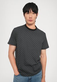 T-shirt noir à manches courtes avec un motif géométrique gris répété, col rond avec bord noir, coupe décontractée et texture de tissu douce.