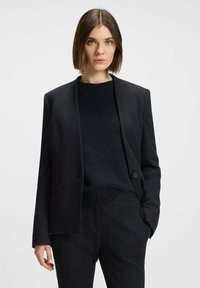 Sort herringbone blazer med spidse lapels og enkelt knaplukning, parret med en mørk strikket sweater og tætsiddende bukser.
