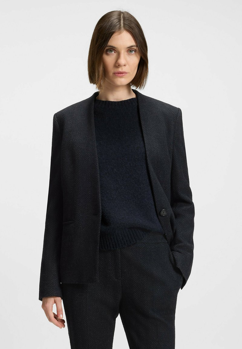 Sort herringbone blazer med spidse lapels og enkelt knaplukning, parret med en mørk strikket sweater og tætsiddende bukser.