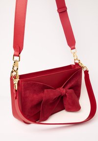 Borsa a mano rossa con un corpo in pelle lucida, un fiocco in camoscio e una tracolla removibile con hardware dorato. Forma rettangolare con texture liscia.