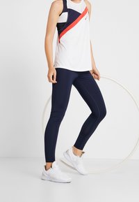 Camiseta de tirantes deportiva con cuerpo blanco y acentos diagonales en azul marino y rojo; combinada con leggings azul marino y zapatillas deportivas blancas. Tejido suave, diseño ajustado.