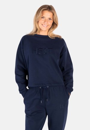 Kvinne som smiler, iført marineblå langermet genser og matchende bukser med "HÉST" preget på begge plaggene, hendene i lommene.