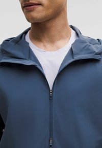 Blauwe zip-up jas met capuchon, gemaakt van glad, lichtgewicht materiaal. Heeft een wit T-shirt eronder en een metallic ritsdetail.