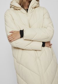 Person wearing a cream-colored quilted puffer jacket with hood, black sweater sleeves visible, arms crossed against a plain background.