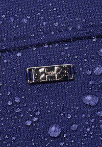 Voděodolná fialová látka s viditelnými kapkami a lesklým stříbrným logem. Textura vypadá jako pletenina s jednotným vzorem.