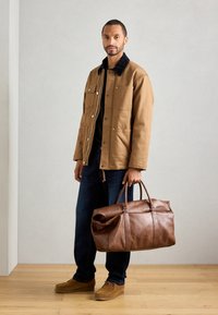 Hombre con chaqueta beige y jeans oscuros sosteniendo un gran bolso de cuero marrón, de pie sobre un suelo de madera contra una pared clara.
