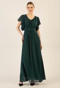 Langes, fließendes grünes Kleid aus leichtem Stoff; hat Rüschenschößchen und eine betonte Taille, mit einer glatten, drapierten Textur.