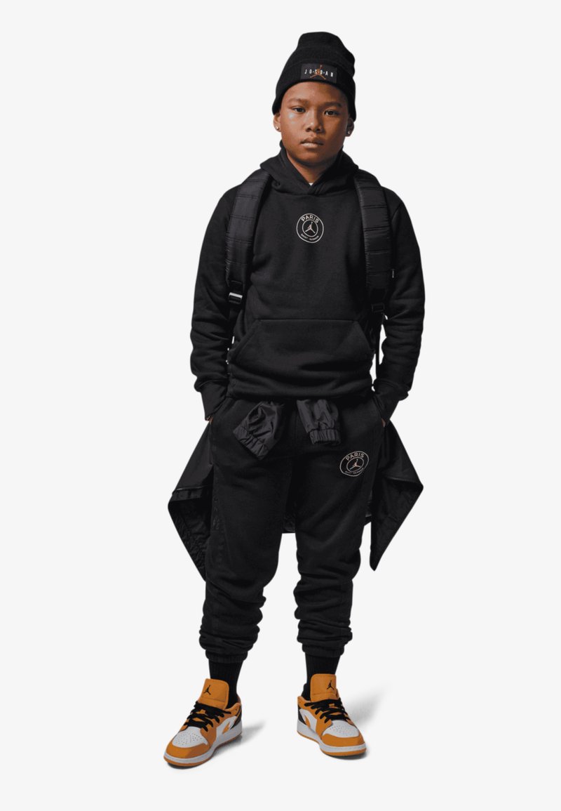 Czarna bluza z kapturem i dresy z logo, zestawione z czapką beanie w kolorze czarnym oraz trampkami z pomarańczowymi akcentami. Wyposażony w czarny plecak.