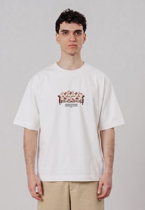 Ung mand med kort krøllet hår iført en hvid oversized t-shirt med et blomsterstolmotiv og beige bukser mod en ensfarvet baggrund.