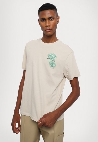 Beige cotton t-shirt featuring a green textured floral appliqué on the chest, short sleeves, and a crew neck design.