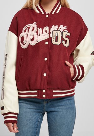 Vrouw draagt een bordeauxrode en crème varsityjas met patches van "Bronx," "05," en "FUBU," gecombineerd met een blauwe spijkerbroek.