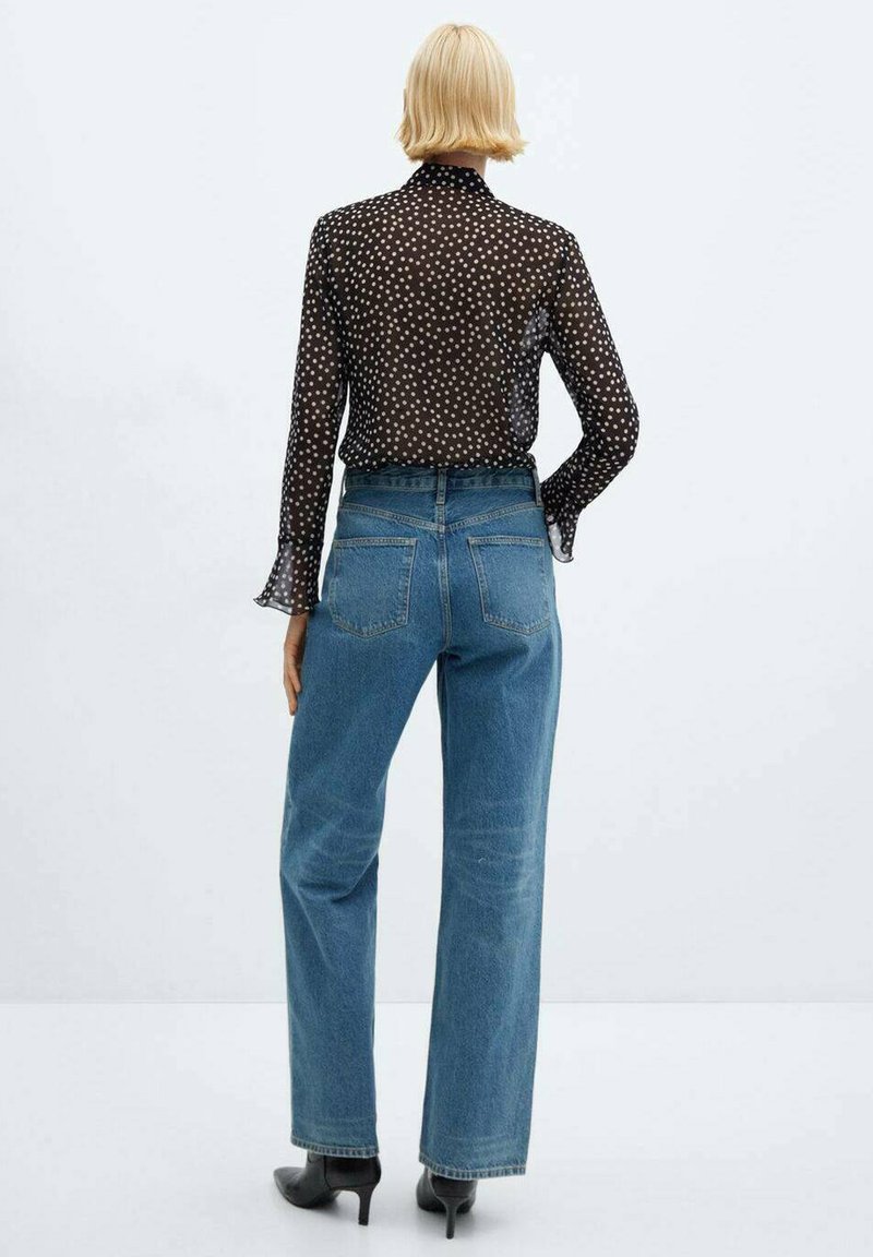 Schwarze transparente Bluse mit weißen Punkten, langen Ärmeln und ausladenden Manschetten, kombiniert mit hochtaillierten, hellblauen weiten Jeans. Schwarze Stiefeletten mit Absatz.