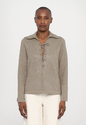 Lace-up, long-sleeve top in a textured, metallic gold fabric with a collar and open neckline, paired with cream-colored pants.