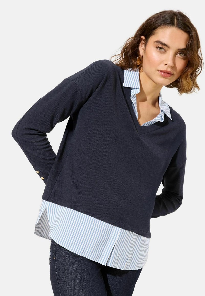 Pull marine à col en V, superposé à une chemise à col rayée bleu clair et blanc. Manches longues avec détails de boutons dorés aux poignets.