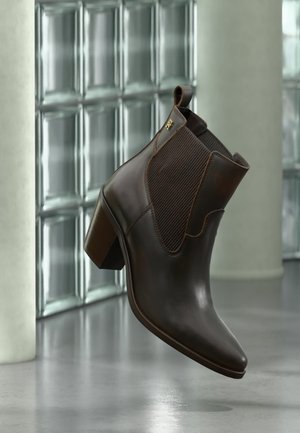 Bottine en cuir marron foncé avec talon bloc et panneaux latéraux élastiques, présentée devant un mur moderne en blocs de verre et un sol en béton.
