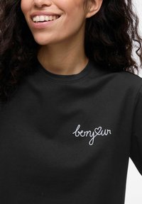 T-shirt nera con ricamo bianco che presenta la parola "bonjour" e un cuore. Il tessuto sembra morbido, con un classico scollo rotondo e maniche corte.