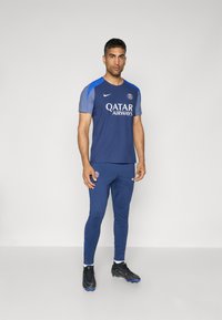 Temno modra atletska trenirka z majico kratkih rokavov, ki ima modre detajle in logotip Qatar Airways, v kombinaciji z ujemajočimi se ozkimi hlačami.