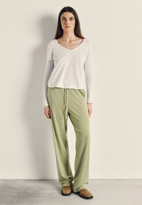 Žena stojící opřená o hladkou zeď, oblečená v bílém dlouhém tričku s výstřihem do V, volných světle zelených kalhotech se šňůrkou a hnědých botách.