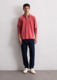 Langermet korallfarget polo skjorte med krage, kombinert med mørkeblå bukser og hvite sneakers. Jevn tekstur, enkelt design.