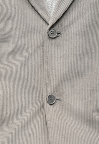 Šedá kordurová vesta s ribovanou texturou, dvěma knoflíky a výstřihem do V. Látka vypadá měkce a má jemný lesk.