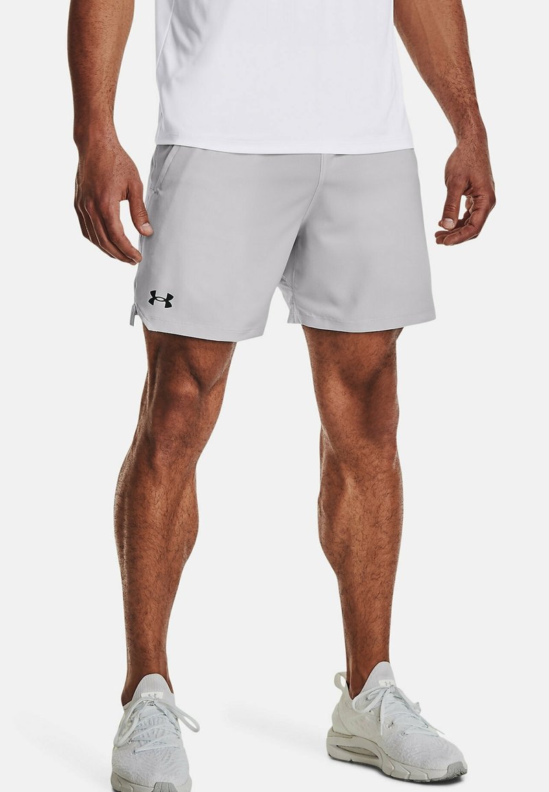 Homme portant un short de sport gris clair Under Armour, des baskets blanches et un t-shirt blanc, debout les jambes légèrement écartées sur fond blanc.