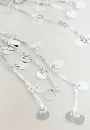 Sudraba ķēdes kaklarota ar maziem apaļiem monētu līdzīgiem piekariņiem uz gaiši neitrāla fona, ar gareniskiem posmiem un spīdīgām diskām.