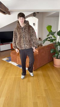 Veste zippée en fausse fourrure imprimé léopard avec capuche, associée à un pantalon noir et des chaussures argentées. Cadre intérieur avec sol en bois et plantes.