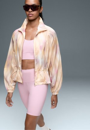 Femme marchant avec une veste tie-dye pastel oversize, un soutien-gorge de sport rose et un cuissard de vélo, des lunettes de soleil foncées et des chaussettes blanches sur un fond uni.