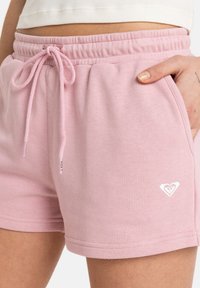Světle růžové bavlněné šortky s elastickým pasem, detaily na šňůrce, bočními kapsami a bílým logem na levé dolní noze.