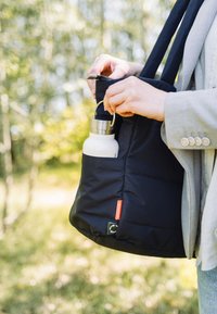 Svart vadderad tygväska med långa remmar, med en sidoficka som håller en vit vattenflaska med lock i rostfritt stål.