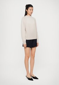 Maglione beige chiaro con maniche raglan, realizzato in un materiale morbido. Abbinato a pantaloni corti blu scuro e ballerine nere a punta.