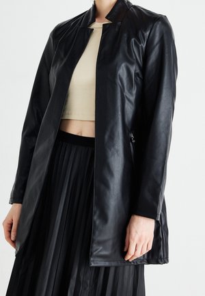 Vrouw draagt een lange zwarte jas van nepleer over een beige cropped top en een zwarte geplooide rok, staand tegen een witte achtergrond.