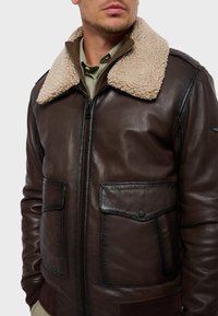 Chaqueta de cuero marrón con cuello de borreguito beige, cierre frontal con cremallera y dos bolsillos de solapa con botones a presión.