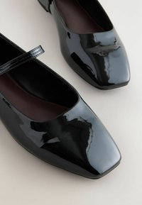 Zwarte lakleren schoenen met een vierkante neus en een dunne band over de wreef. De binnenkant is van een contrasterend donkerbruin materiaal.