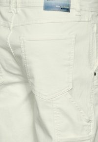 Pantalones de denim blancos con un bolsillo trasero de forma rectangular, detalles de costura sutiles y una etiqueta con la leyenda "RELÁJATE Y DESCONÉCTATE".