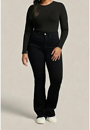 Frau trägt ein eng anliegendes schwarzes Langarmshirt, hoch taillierte schwarze Hose und weiße Schuhe vor neutralem Hintergrund.